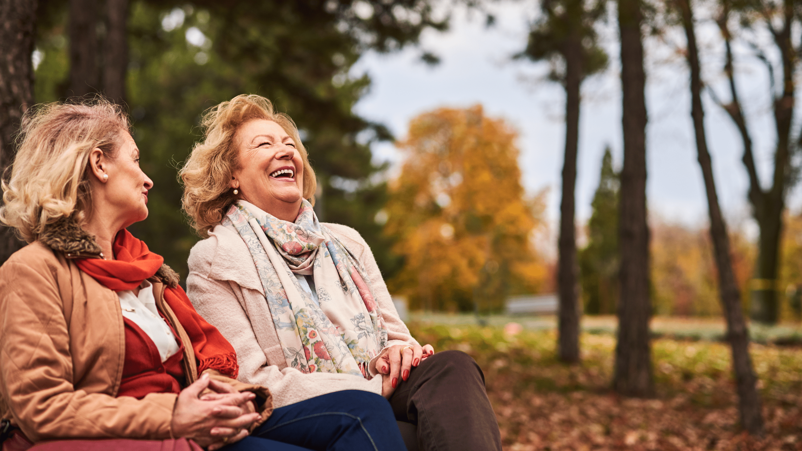 Op de afbeelding zie je twee dames van rond de 60. Ze zitten samen gezellig op een herfstachtige dag op een bankje te kletsen en te lachen.